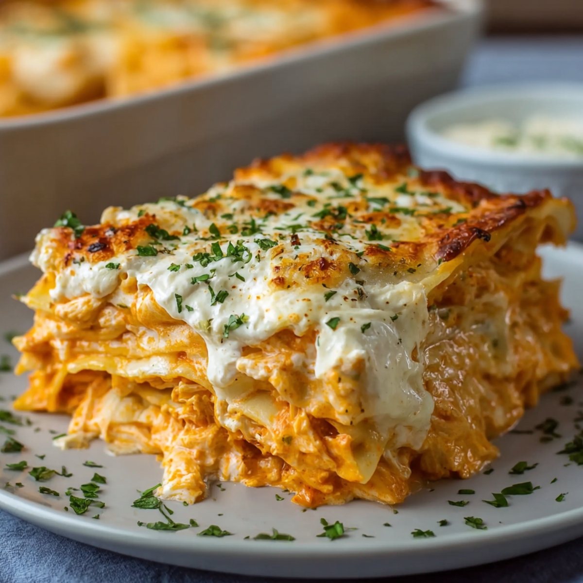 Buffalo chicken lasagna slice with creamy shredded chicken, melted cheese, and golden-browned top, garnished with fresh parsley on a dark plate.