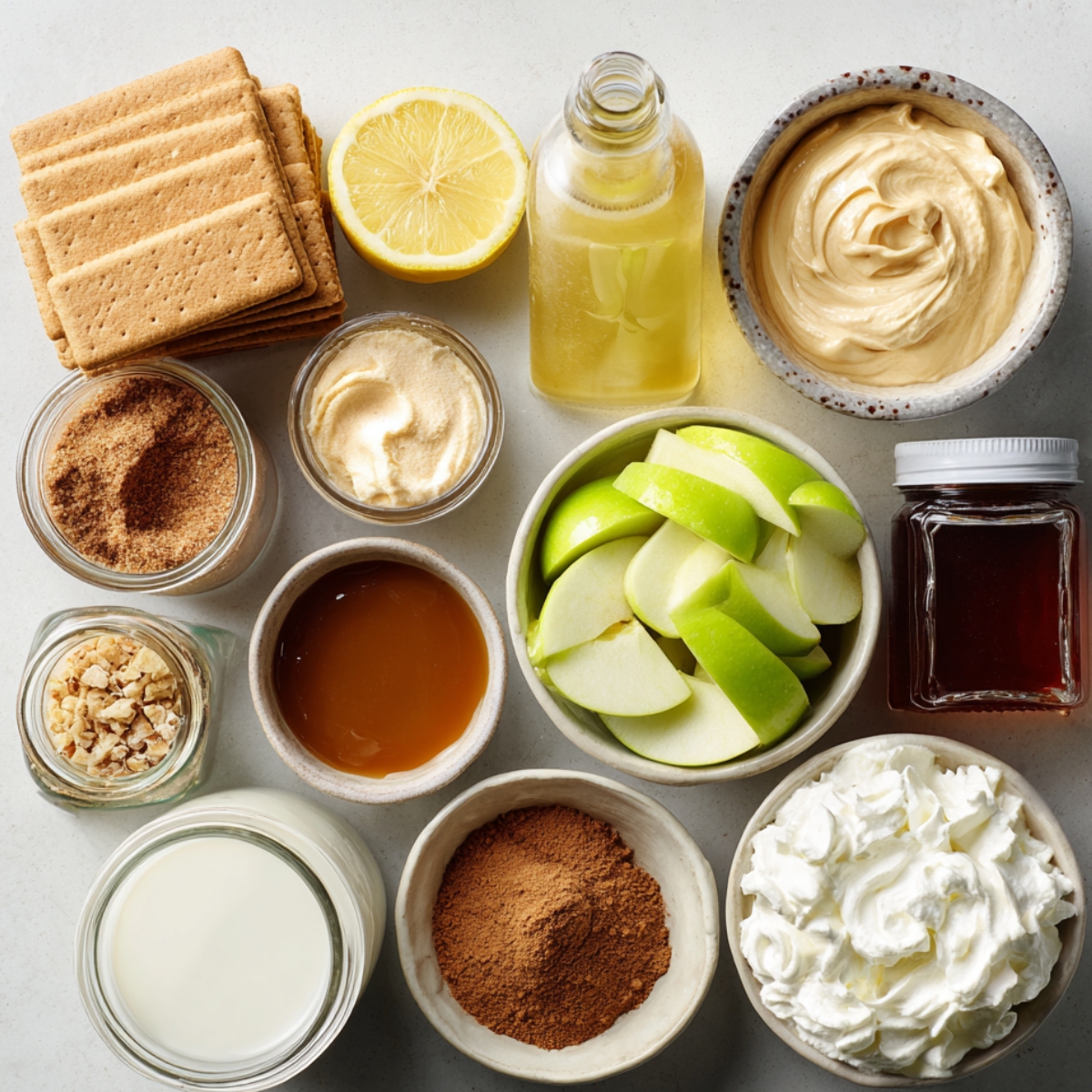 Ingredients for Caramel Apple Eclair Cake, including graham crackers, lemon, vanilla extract, whipped cream, caramel sauce, sliced apples, brown sugar, cinnamon, walnuts, milk, and maple syrup, arranged on a countertop.