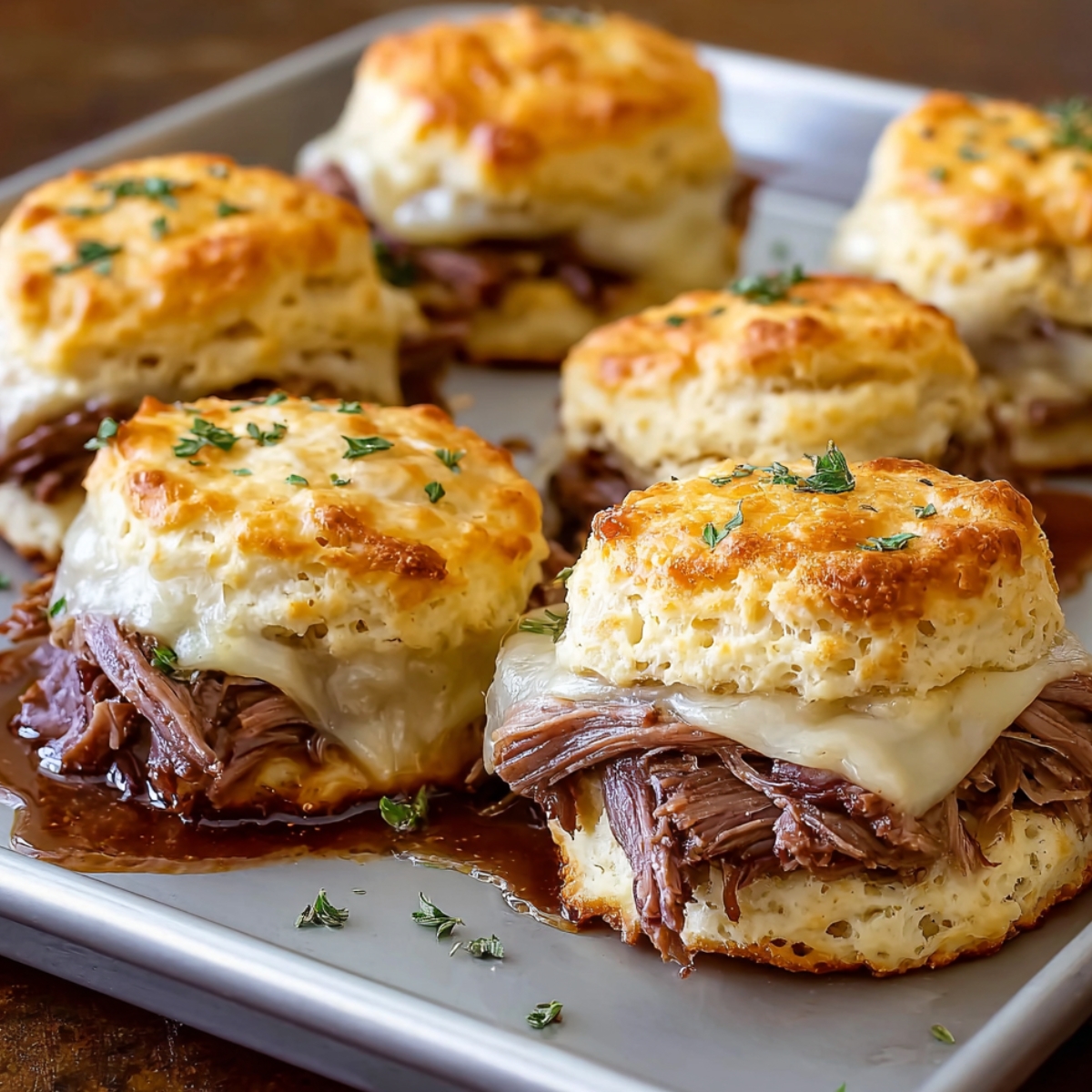 Golden French Dip Biscuits filled with shredded roast beef and melted cheese on a baking sheet, topped with herbs and warm au jus, cozy homemade look.