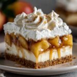 Homemade Caramel Apple Eclair Cake with a graham cracker crust, creamy filling, caramelized apples, and whipped cream topped with cinnamon, served on a beige plate with a blurred red apple in the background.