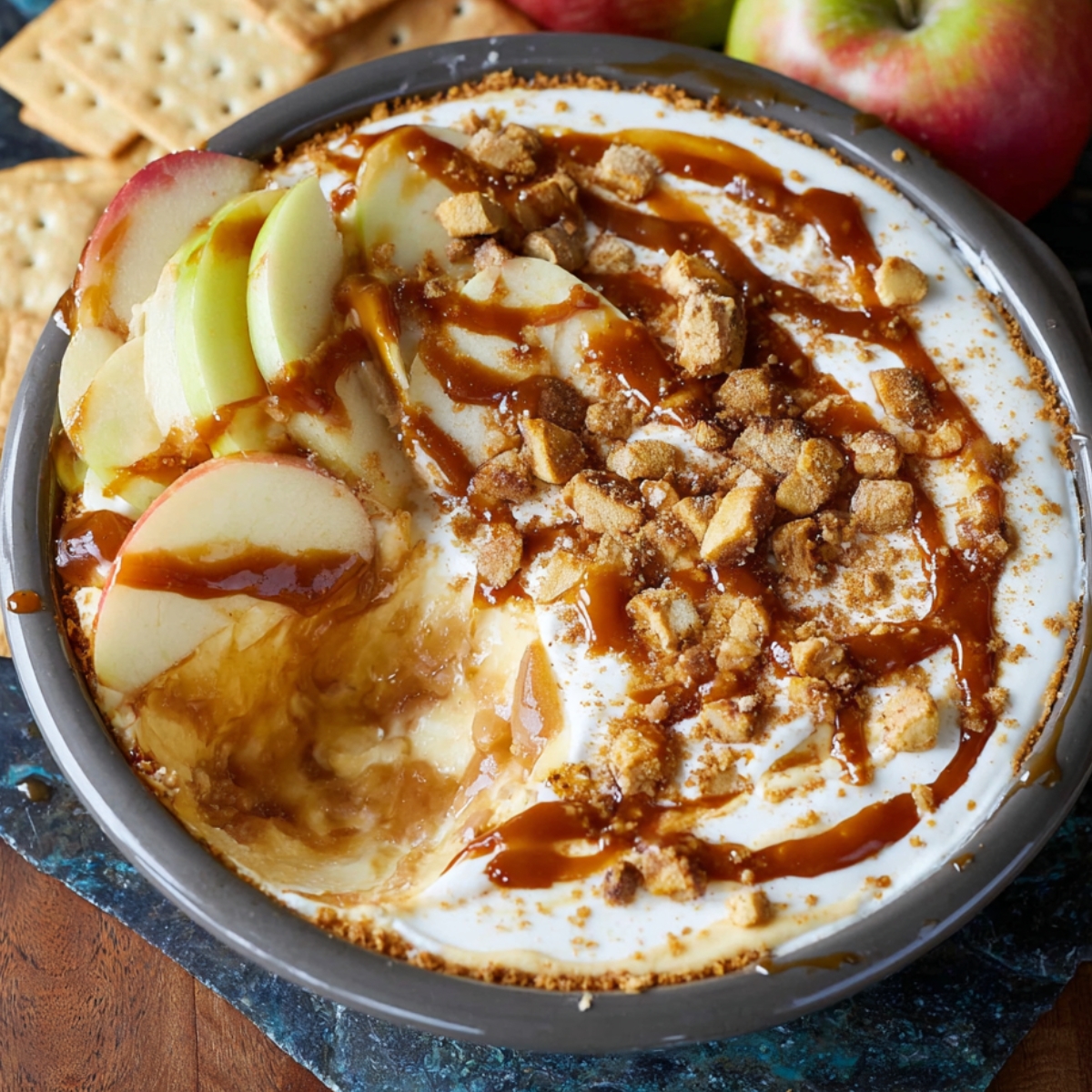 Homemade caramel apple cheesecake dip topped with sliced apples, caramel drizzle, and crumbled graham crackers in a round dish, with fresh apples and crackers in the background.