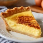 Close-up of a homemade egg custard pie slice on a vintage white and gray patterned plate, showing a smooth, creamy yellow custard filling topped with a golden-brown nutmeg-speckled surface and a flaky, buttery pie crust edge, softly lit on a wooden table with a blurred background for a warm, inviting kitchen feel.
