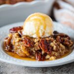 Homemade Pecan Pie Cobbler topped with a scoop of melting vanilla ice cream, featuring caramelized pecans and a gooey brown sugar filling on a white dessert plate.