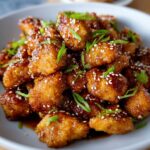 Honey Garlic Chicken Bites coated in a sticky glaze, topped with sesame seeds and sliced green onions on a plate.