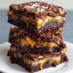 Stack of homemade Outrageous Snickers Brownies with rich chocolate layers, gooey caramel filling, and a glossy drizzle on top, sprinkled with flaky sea salt, served on a white plate for a delicious, real, and freshly baked look.