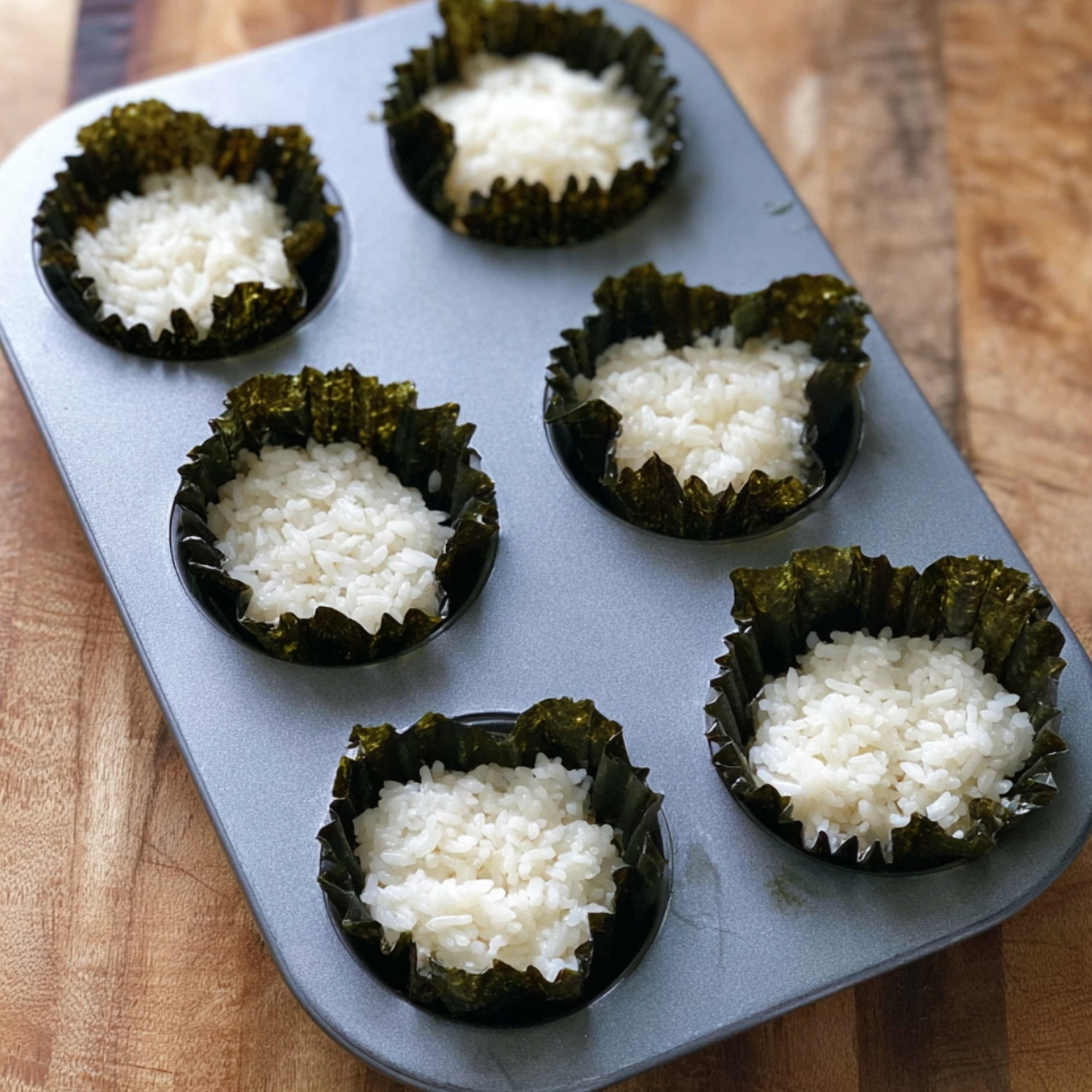 Place one nori square into each muffin cup. Add 2 tablespoons of sushi rice on top of each nori sheet, then use your fingers or the back of a spoon to press the rice down gently. You want it to stick to the nori and form a little base.