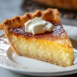 Slice of homemade buttermilk pie with a golden, flaky crust, creamy filling, and topped with a dollop of whipped cream, served on a white plate.