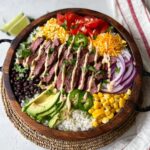 A homemade Cilantro Lime Steak Bowls filled with sliced grilled steak, rice, black beans, avocado, corn, tomatoes, red onion, jalapeños, shredded cheese, and a creamy drizzle, all arranged neatly in a rustic wooden bowl.