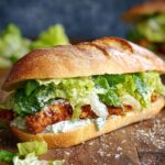 A Homemade Crispy Chicken Caesar Sandwich with fresh lettuce, creamy dressing, and parmesan cheese on a toasted baguette, served on a wooden surface.