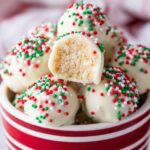 Homemade Sugar Cookie Truffles dipped in white chocolate and decorated with red, green, and white sprinkles, with one truffle halved to reveal its soft, crumbly center.