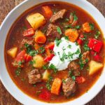 A hearty bowl of homemade Hungarian goulash filled with tender beef, potatoes, carrots, and red peppers, topped with a dollop of sour cream and fresh parsley.