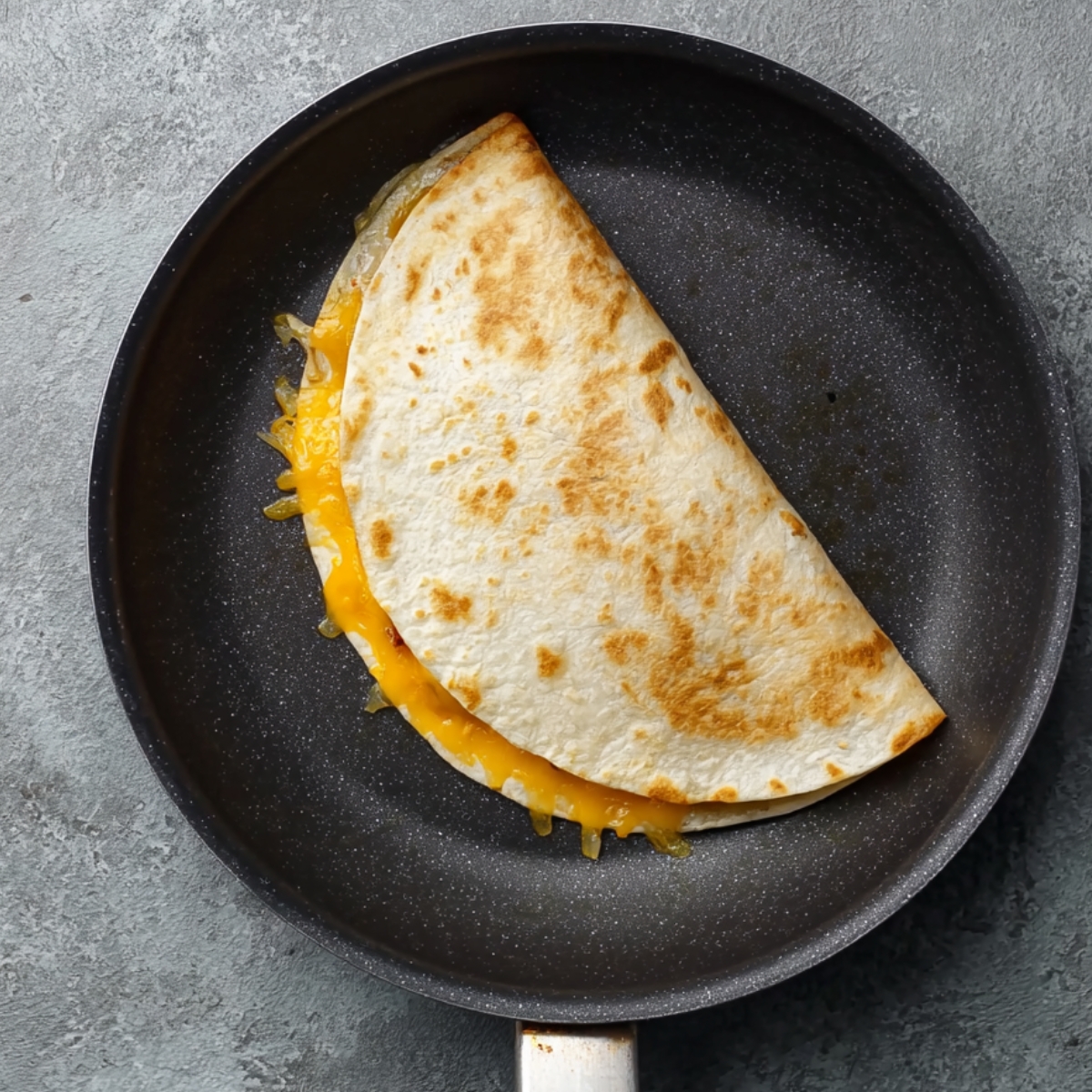 Tortilla with cheese, ground beef, and pickles on a skillet, ready to be folded into a quesadilla.