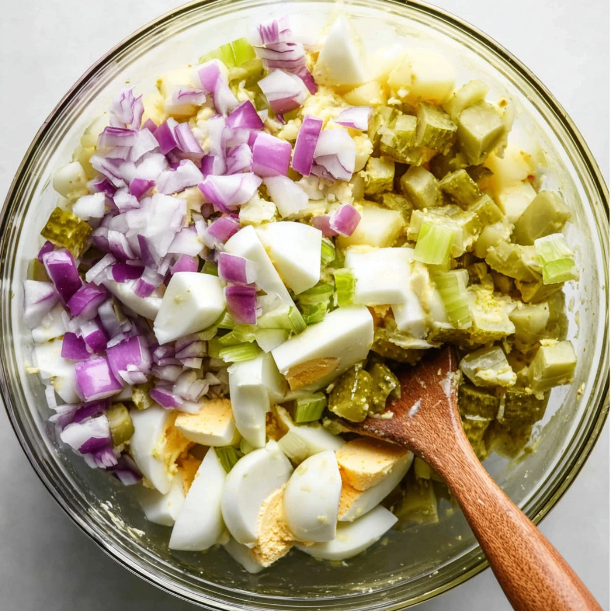 A mixing bowl containing the key ingredients for potato salad – diced potatoes, hard-boiled eggs, chopped celery, pickles, and onions. A wooden spatula is ready for mixing.