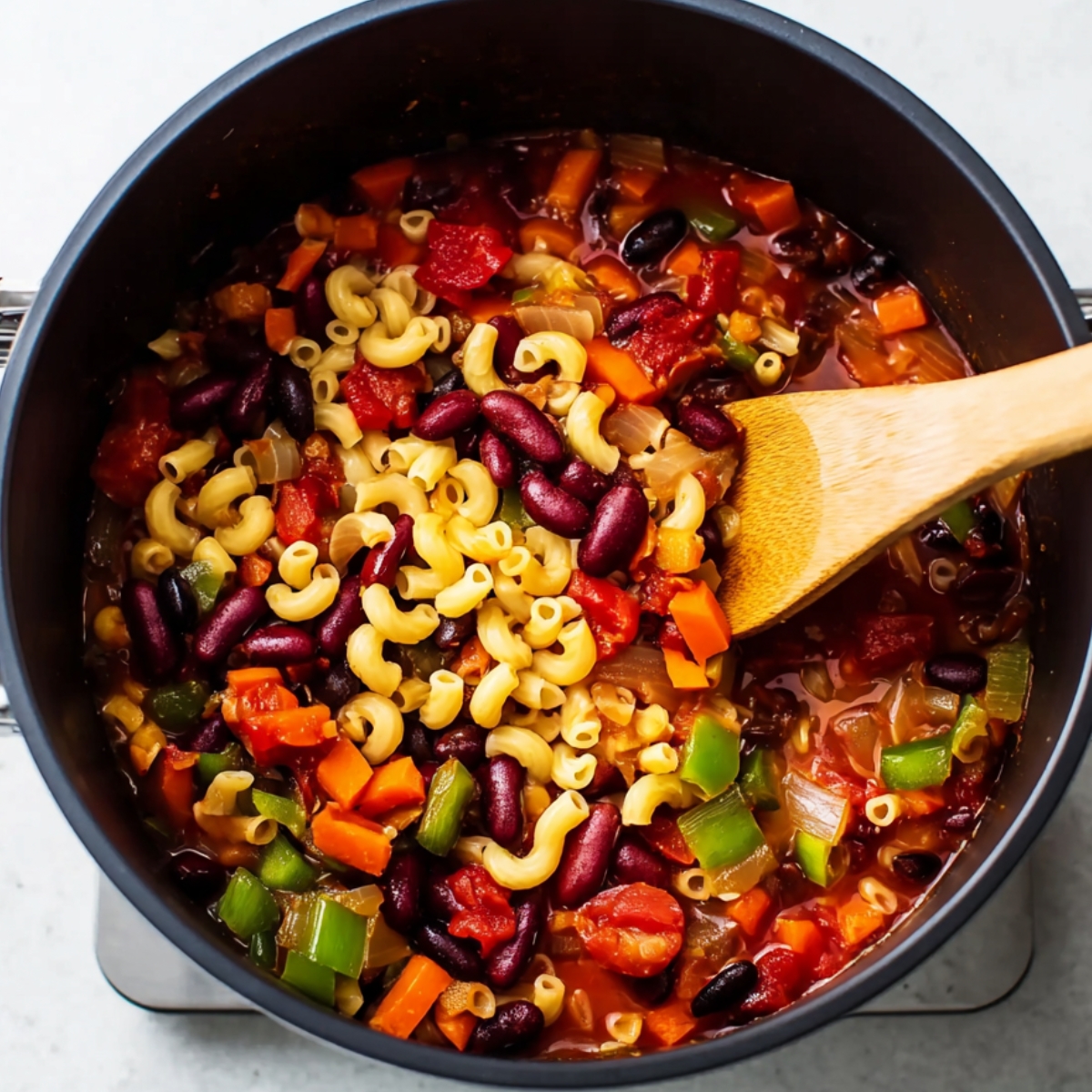 Vegetarian chili mac cooking in a pot with elbow pasta, kidney beans, black beans, tomatoes, carrots, and bell peppers being stirred with a wooden spoon