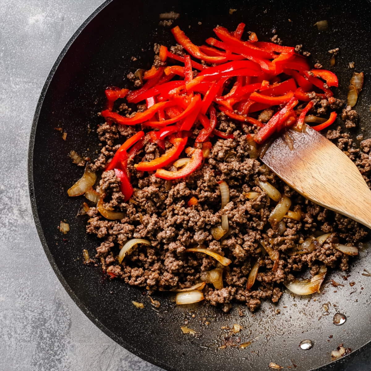 A wok with browned ground beef and red bell pepper slices being stir-fried, with a wooden spoon mixing the ingredients, ready to be seasoned and served.