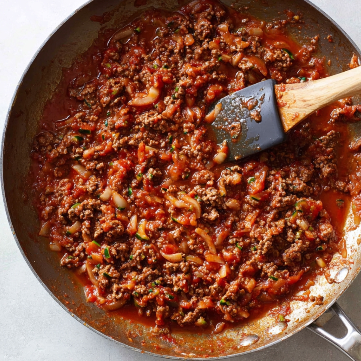 A skillet with cooked ground beef, onions, and tomatoes simmering in a savory tomato sauce. The beef is browned and crumbled, and the mixture is thick and rich, perfect for layering in a casserole.
