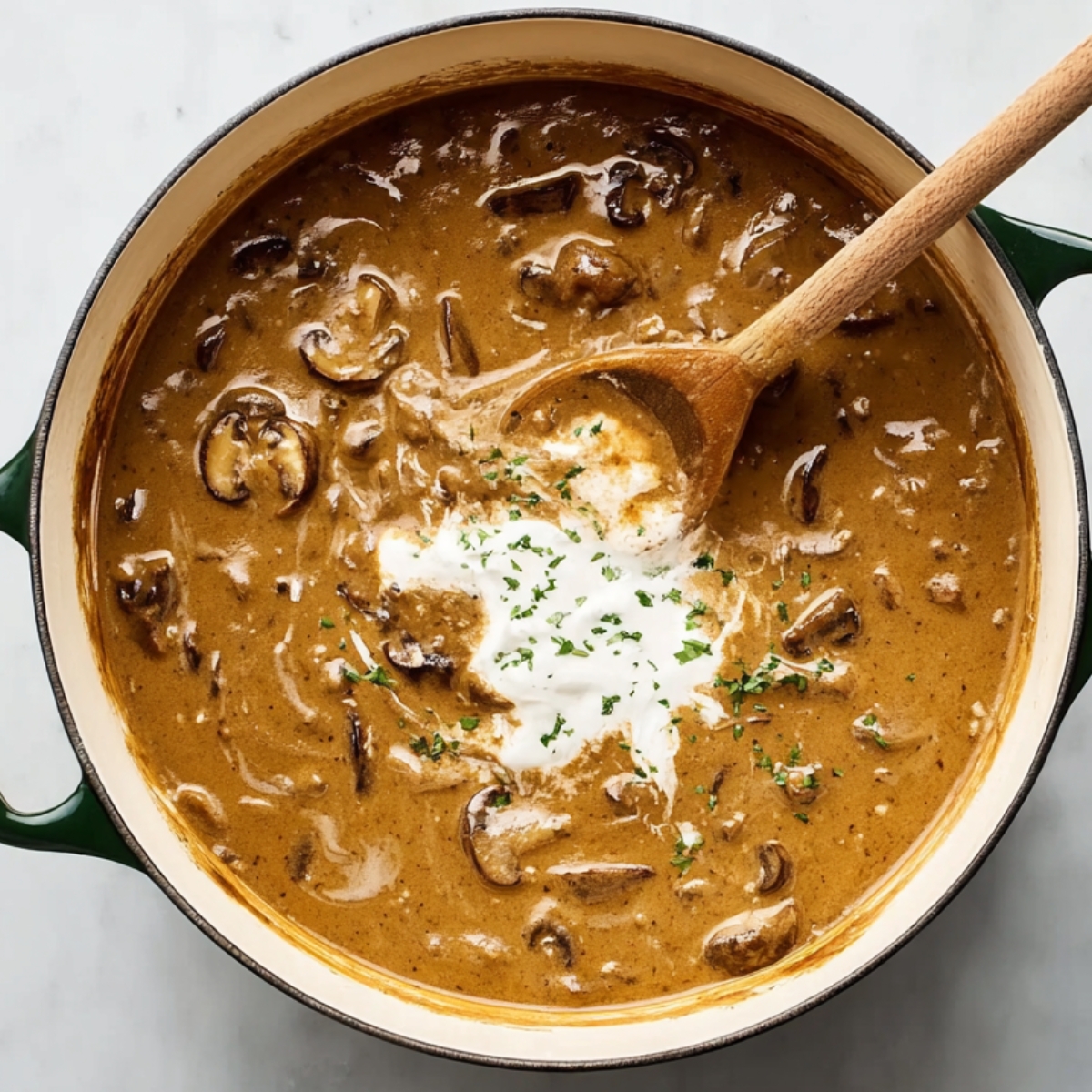 Creamy Hungarian mushroom soup in a green pot with a wooden spoon stirring in sour cream and fresh herbs.