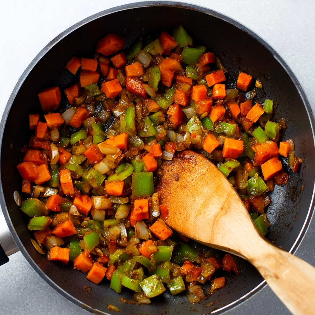 Sautéed carrots, green bell peppers, and onions cooking in a skillet with spices and a wooden spoon