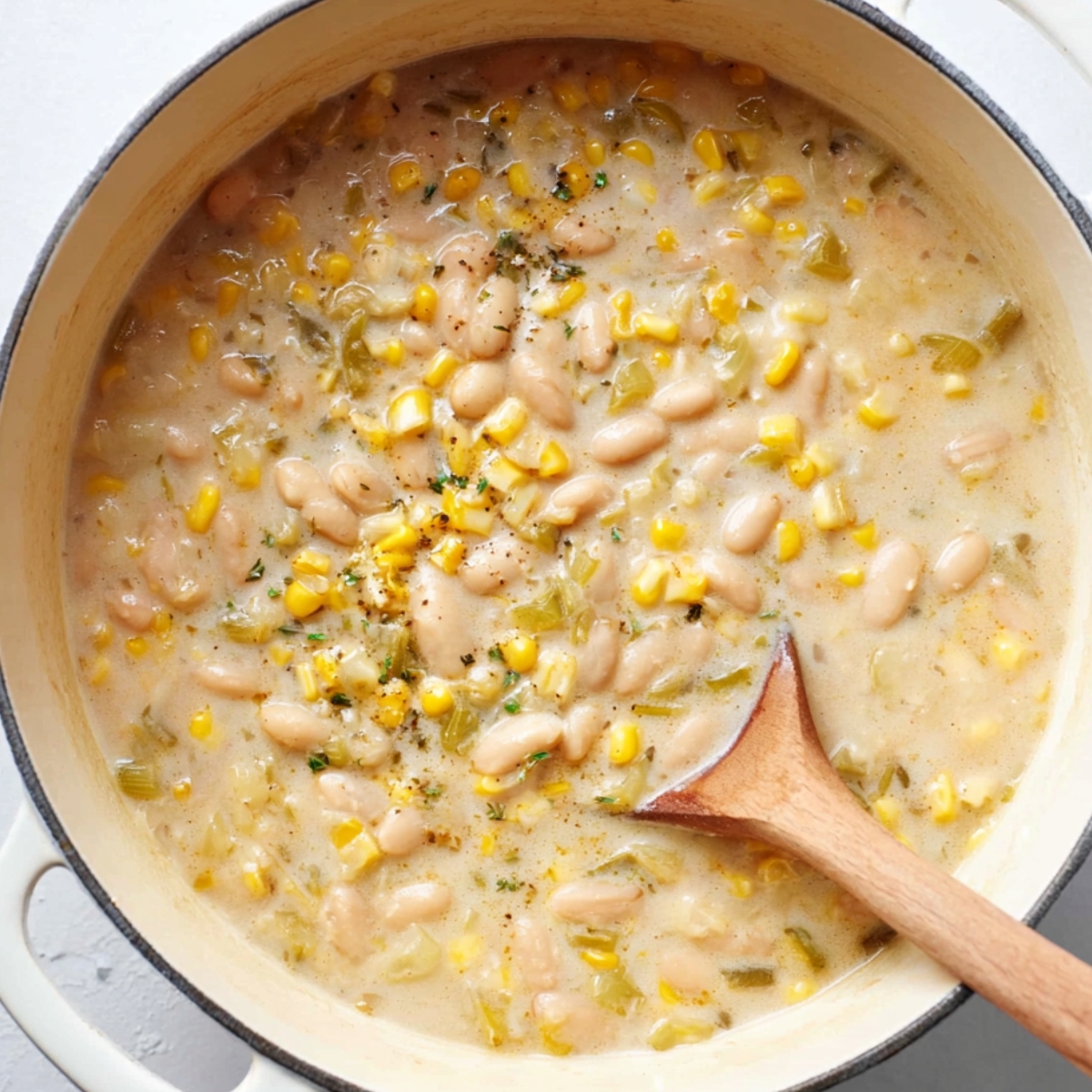 Creamy white bean chicken chili simmering in a pot with corn and herbs, stirred with a wooden spoon.