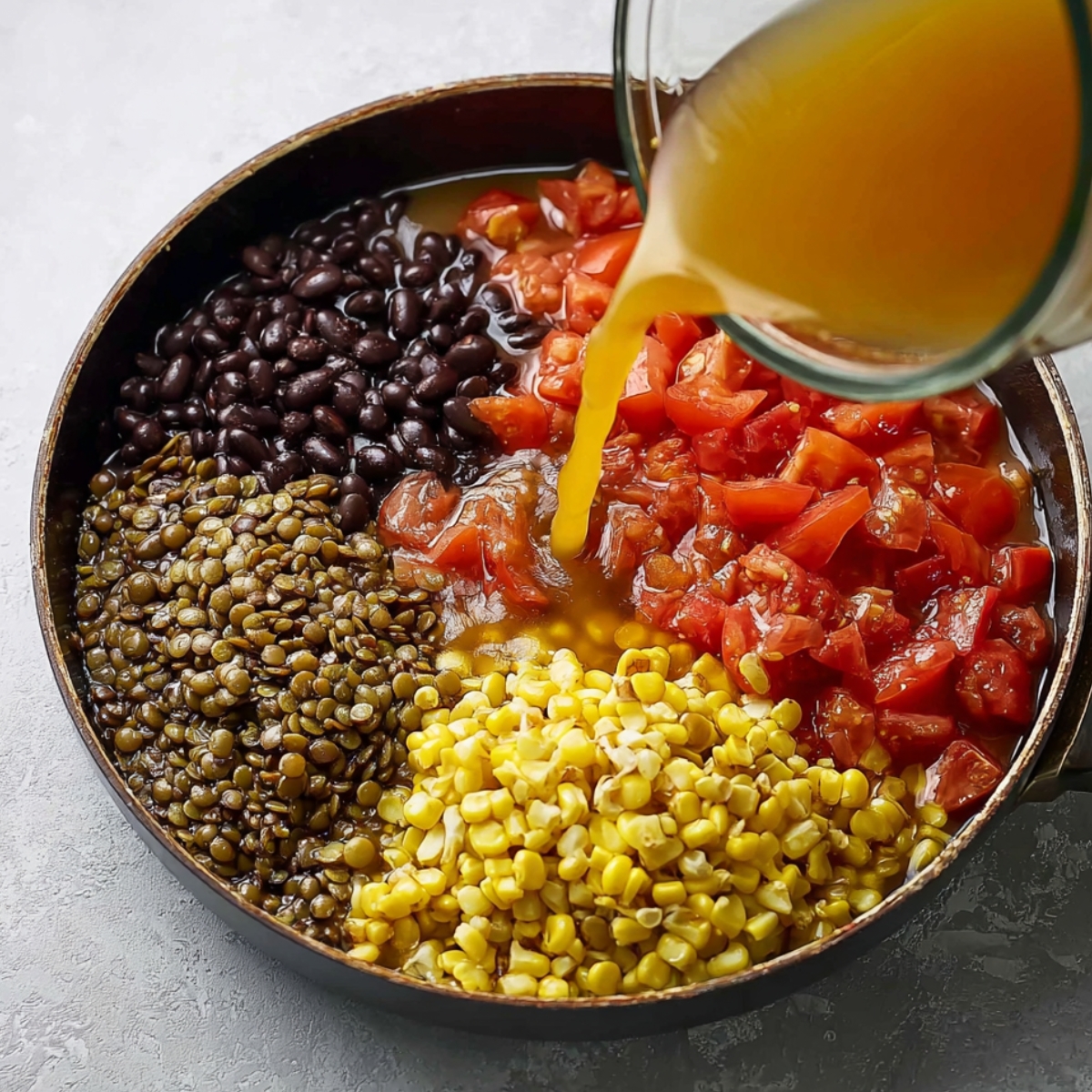 A skillet filled with black beans, corn, diced tomatoes, and lentils, with broth being poured in, preparing the base for a flavorful dish.