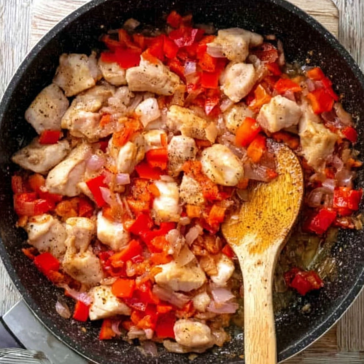 Chicken pieces cooking with red peppers and onions in a skillet, seasoned with black pepper.