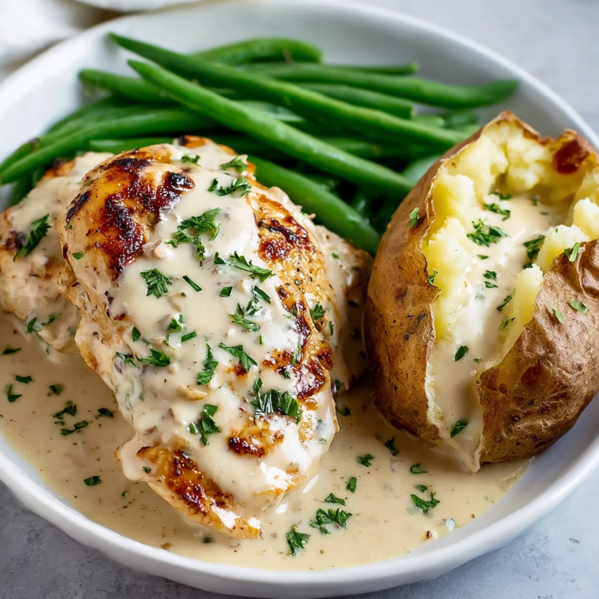 A plate Creamy Ranch Chicken covered in creamy ranch sauce, garnished with chopped parsley. The dish includes green beans and a baked potato with creamy sauce.