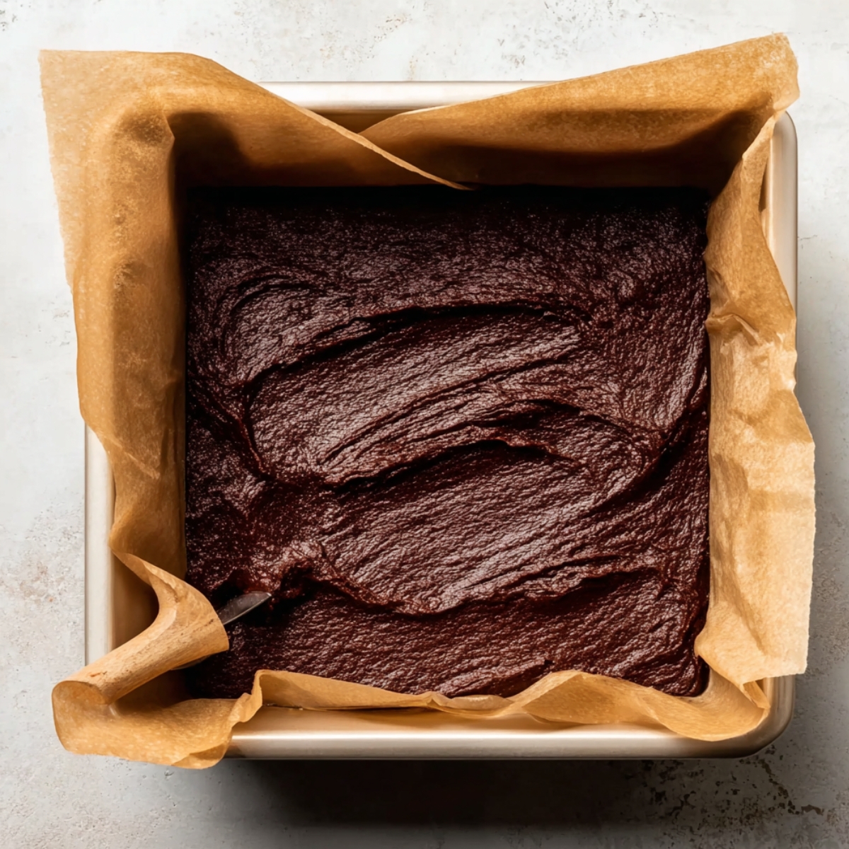 Brownie batter spread evenly into a square baking pan lined with parchment paper.
