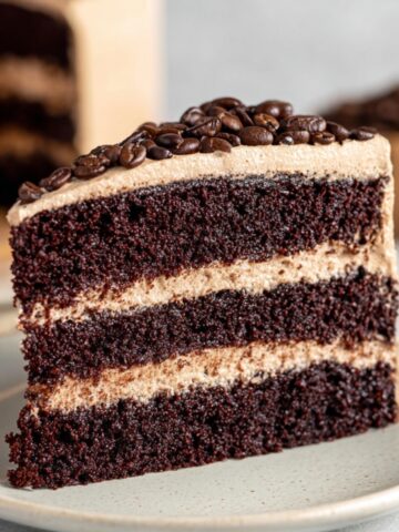 A slice of Coffee Chocolate Cake with layers of moist cake and creamy coffee buttercream frosting, topped with coffee beans, placed on a light plate.