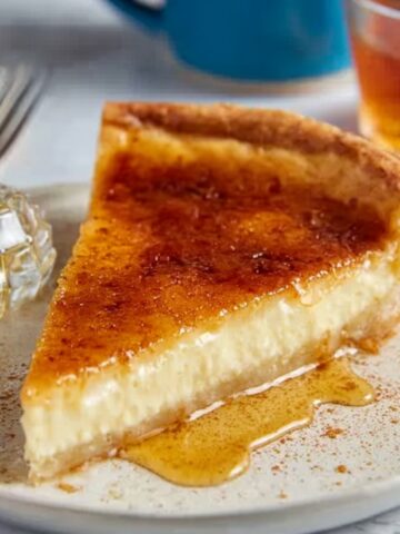 A slice of Greek Honey Pie on a plate, with a golden crust and creamy filling, drizzled with honey. A small piece of honeycomb is placed next to the pie.