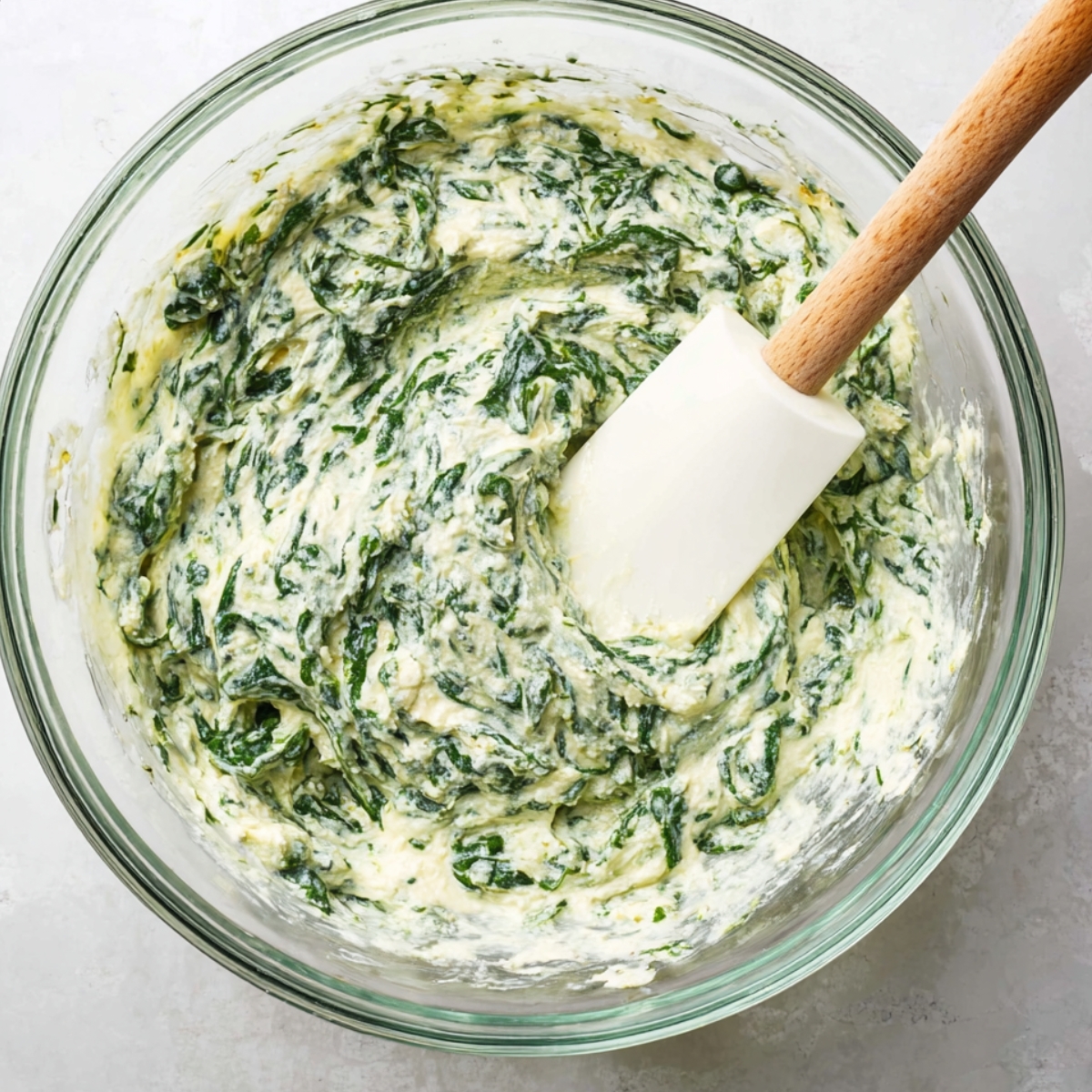 Creamy spinach and ricotta mixture being stirred together in a glass bowl with a spatula.