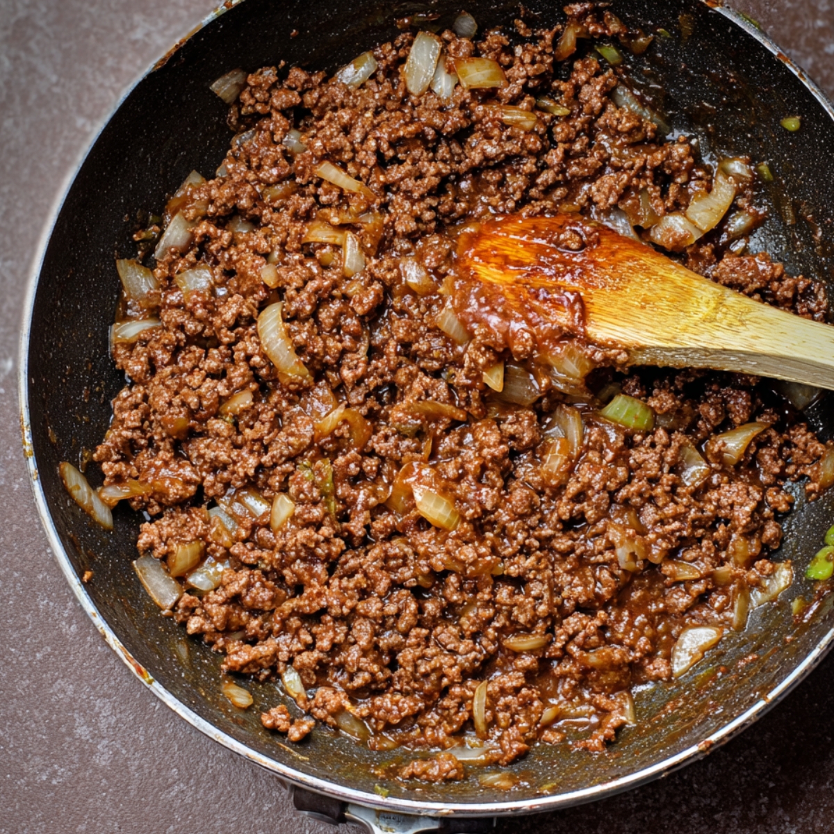 A skillet with seasoned ground beef and onions simmering in a savory sauce, stirred with a wooden spoon.