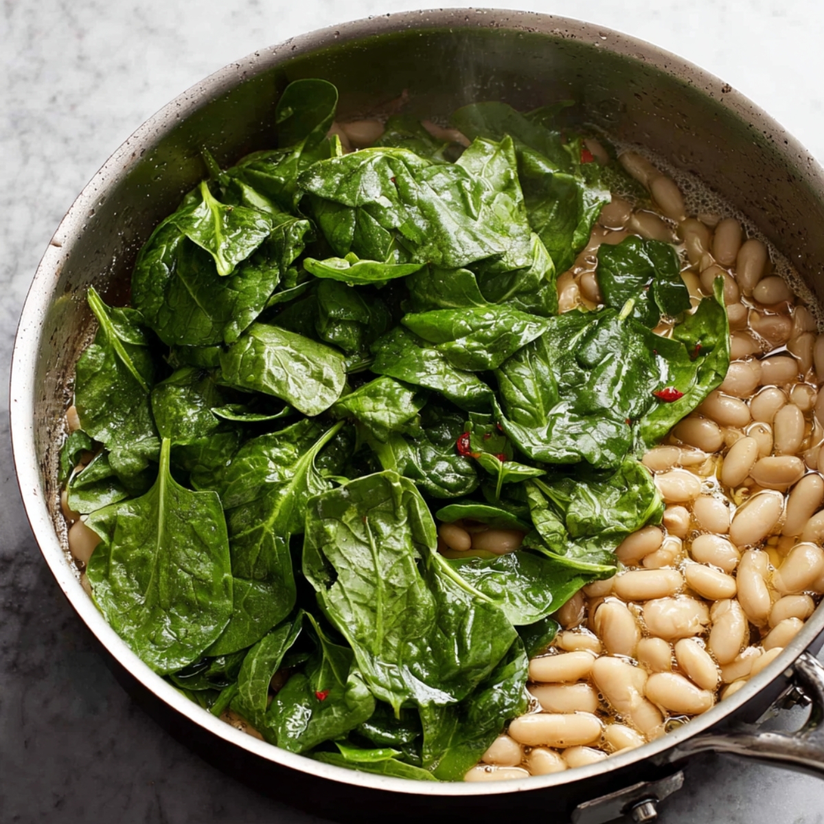 Fresh spinach added to a skillet of cooked white beans, ready to wilt and incorporate into a savory dish.